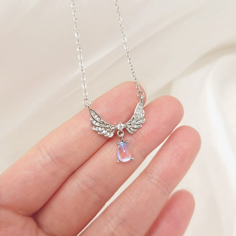 May include: Silver necklace with a teardrop-shaped iridescent gemstone pendant and angel wings with clear crystal accents.