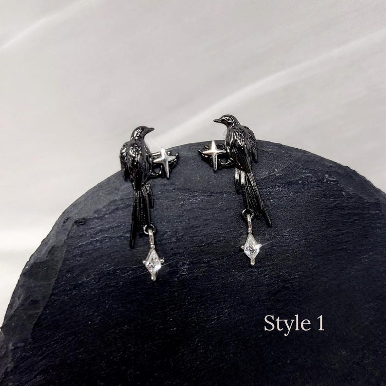May include: A pair of black raven earrings. Each earring features a raven perched on a star, with a dangling, clear, diamond-shaped charm. The earrings are displayed on a dark, textured surface. The text "Style 1" is visible.