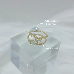 May include: Two stackable, adjustable gold rings. One ring features a rectangular, light green gemstone. The other ring has a decorative, curved design with small, clear gemstones. Text on the image reads: Adjustable. One size.