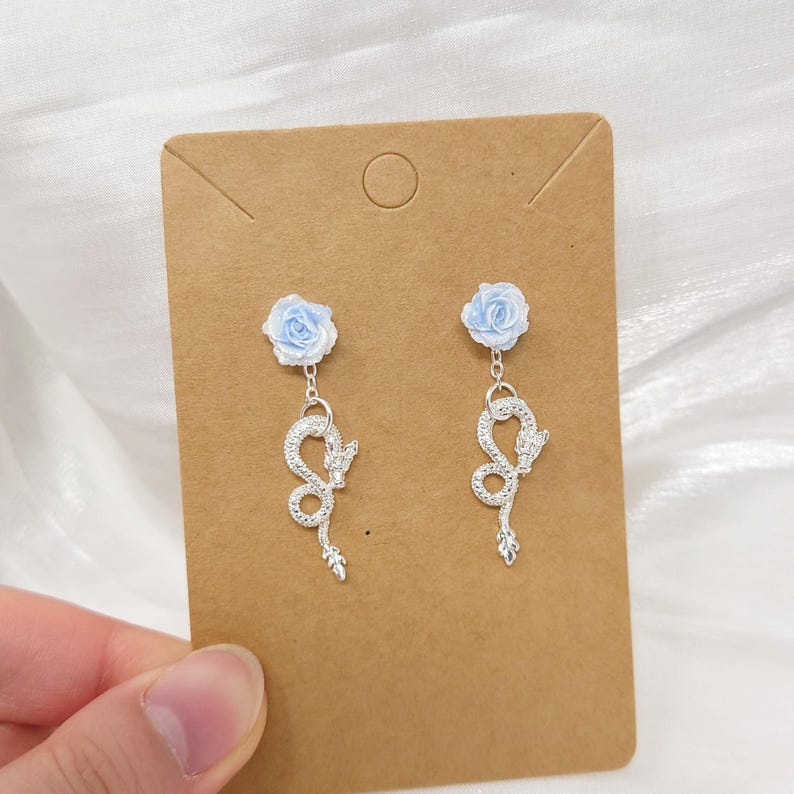 May include: A pair of dangle earrings featuring a light blue rose stud and a silver-colored snake design. The earrings are displayed on a brown card, with a white background.