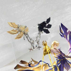 May include: Two pairs of earrings. One pair features gold-colored wings, cream-colored roses, and silver accents. The other pair has black wings and a black rose. Both pairs have a dangling design.
