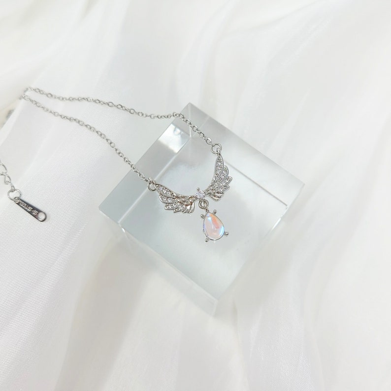 May include: A silver necklace with a teardrop-shaped iridescent stone pendant and a pair of angel wings with clear crystals.