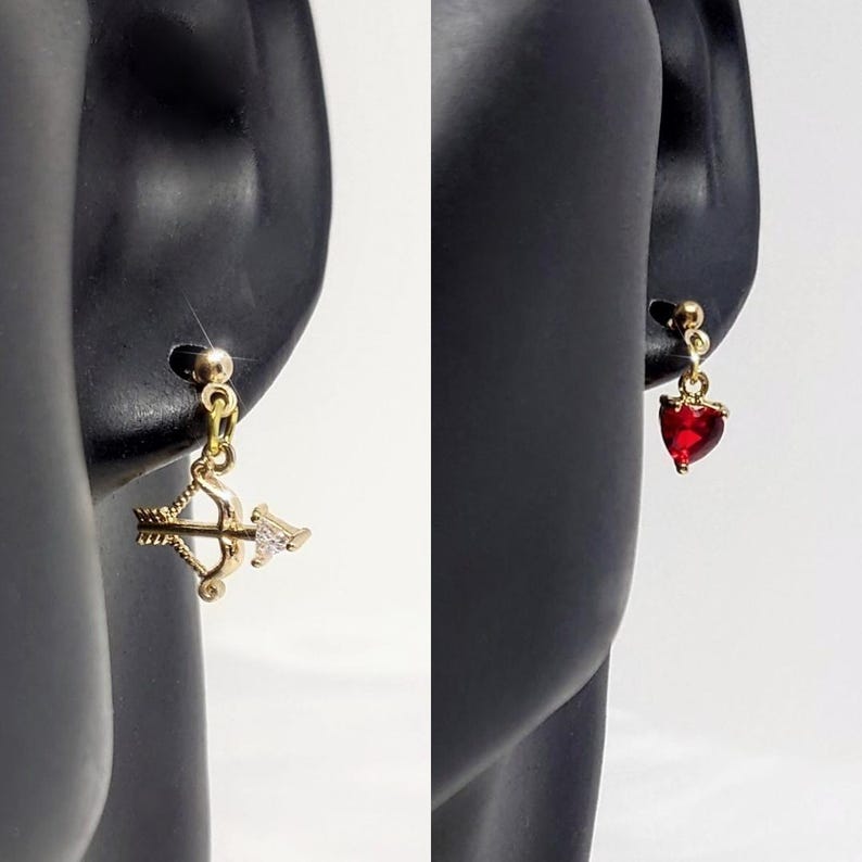 K&ouml;nnte beinhalten: Ein Paar goldene Ohrh&auml;nger. Ein Ohrring hat einen kleinen Bogen- und Pfeil-Anh&auml;nger mit einem klaren Strassstein. Der andere Ohrring hat einen kleinen roten Herz-Anh&auml;nger.