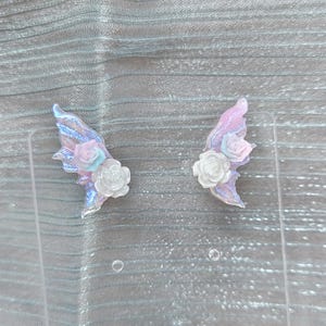 May include: Two iridescent fairy wing earrings with a pink and blue color scheme. Each wing features a white rose and a smaller pink and blue rose. The wings are set against a light blue background.