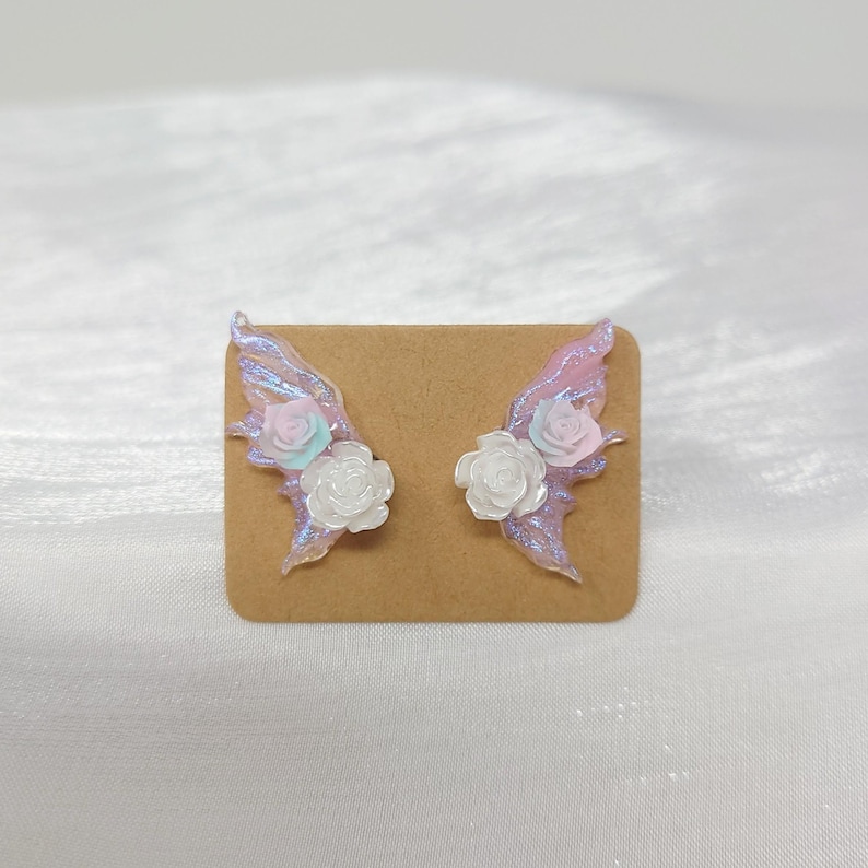 May include: A pair of iridescent fairy wing earrings with white and pastel pink roses. The wings are attached to a brown card. The earrings are a whimsical accessory.