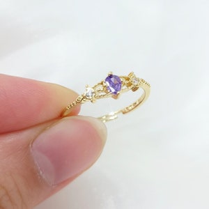 May include: A gold-colored ring featuring a pear-shaped purple gemstone and small clear accent stones. The ring has a delicate, openwork design with a slender band. The ring is held up by a person's fingers.