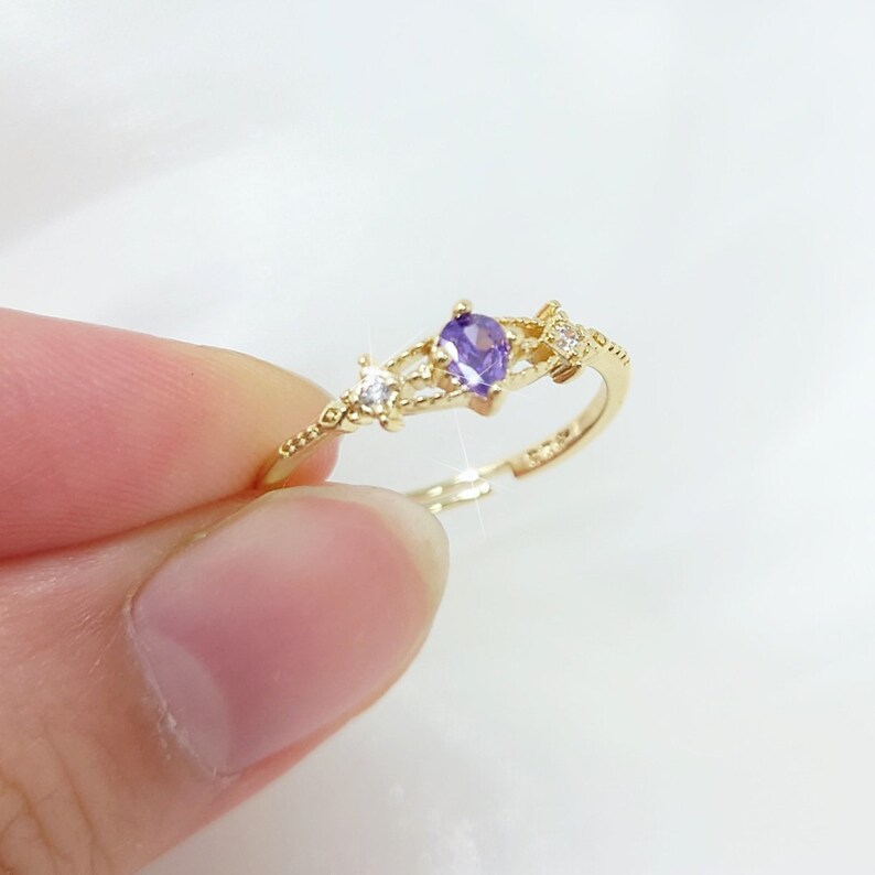 May include: A gold-colored ring featuring a pear-shaped purple gemstone and small clear accent stones. The ring has a delicate, openwork design with a slender band. The ring is held up by a person's fingers.