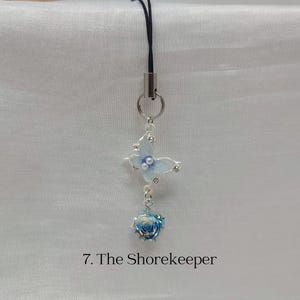 K&ouml;nnte beinhalten: Ein filigraner Anh&auml;nger mit einem hellblauen Schmetterling mit Strassakzenten und einer kleinen blauen Rose. Der Anh&auml;nger h&auml;ngt an einem silbernen Ring und einem schwarzen Band. Der Text "7. The Shorekeeper" steht unten.