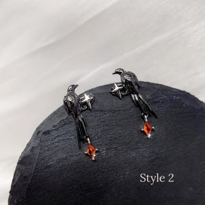 May include: A pair of black raven earrings. Each earring features a raven perched on a star, with a dangling red gem. The earrings are displayed on a dark, textured surface against a white background. The text "Style 2" is visible.