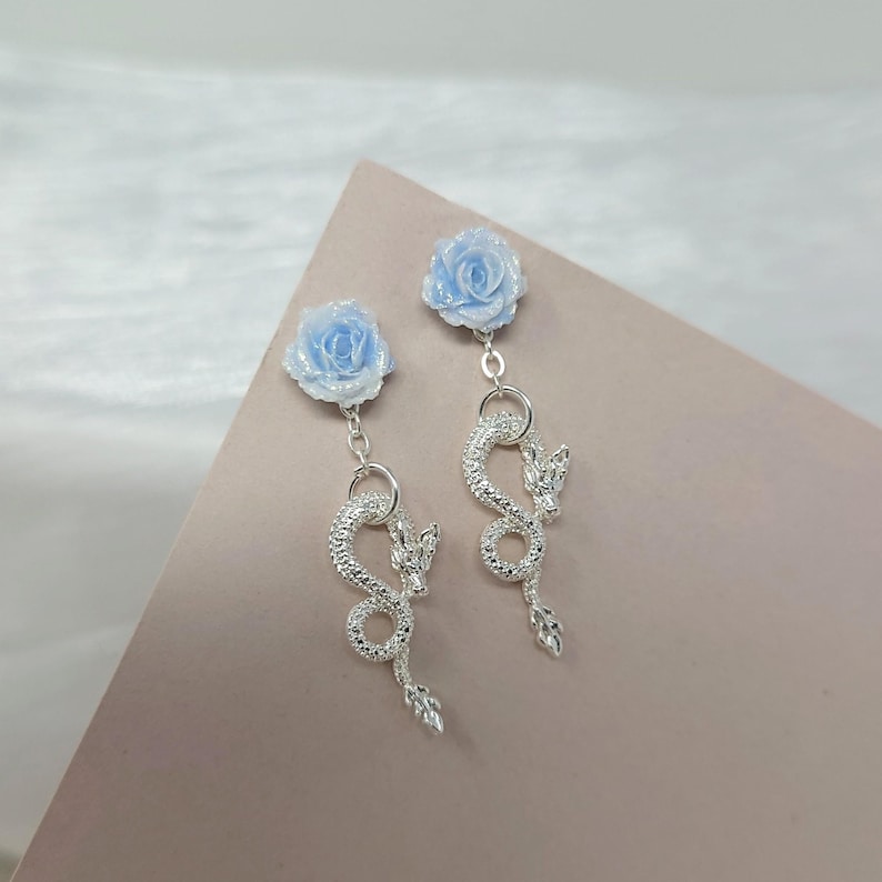 May include: A pair of dangle earrings featuring light blue rose studs. Below the roses, silver dragon pendants with intricate detailing hang from the studs. The earrings are displayed on a pink surface.