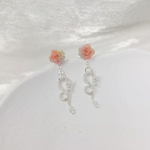 May include: A pair of dangle earrings featuring a rose-shaped stud in shades of pink and yellow. Below the rose, a silver-colored, serpentine design hangs, adding a unique and elegant touch. The earrings are set against a soft, white background.