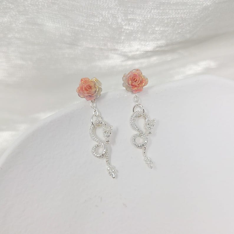 May include: A pair of dangle earrings featuring a rose-shaped stud in shades of pink and yellow. Below the rose, a silver-colored, serpentine design hangs, adding a unique and elegant touch. The earrings are set against a soft, white background.