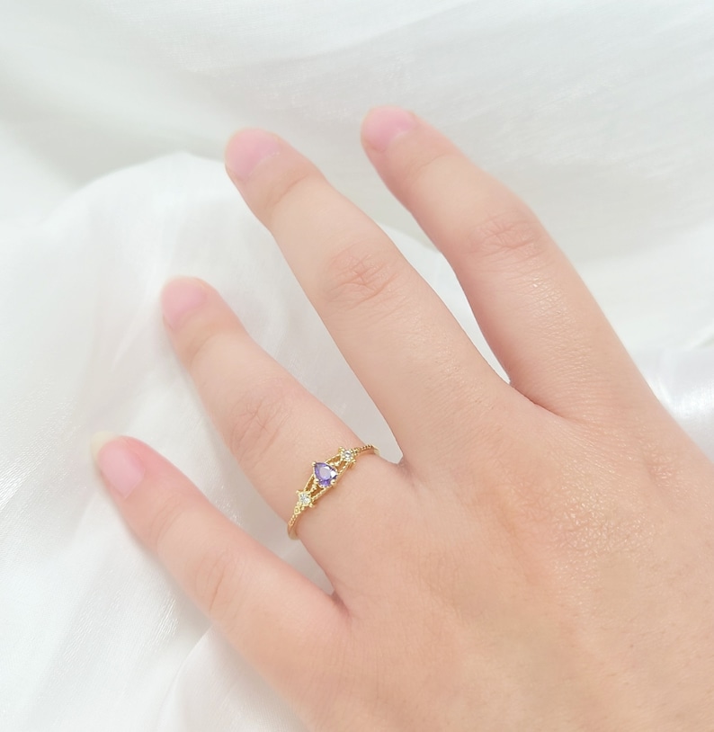 May include: A gold-colored ring featuring a pear-shaped purple gemstone, flanked by two star-shaped clear stones. The ring is set on a delicate band, and is displayed on a white background.
