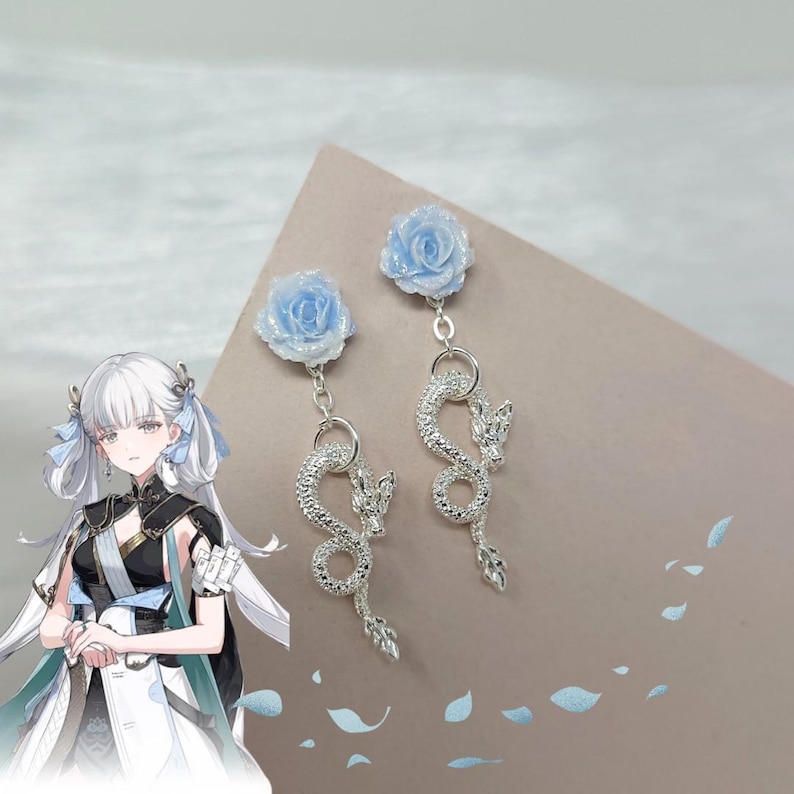 May include: A pair of dangle earrings featuring light blue rose-shaped studs. Below, a silver dragon design hangs from a circular ring. The earrings are displayed on a neutral-toned surface.
