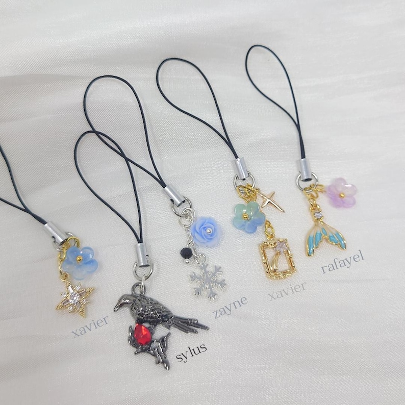 May include: Five phone charms with black cords. Charms include a blue flower with a star, a dark metal raven with a red stone, a blue rose with a snowflake, a blue flower with a cross and frame, and a blue mermaid tail with a purple flower.