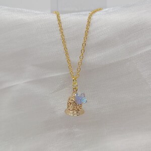 May include: A gold-colored necklace with a pendant shaped like a bell. The bell has intricate cut-out designs and a small, iridescent butterfly charm attached. The chain is delicate, and the background is a soft, white fabric.