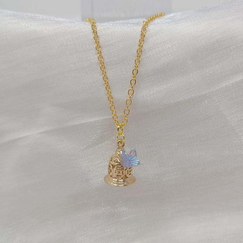May include: A gold-colored necklace with a pendant shaped like a bell. The bell has intricate cut-out designs and a small, iridescent butterfly charm attached. The chain is delicate, and the background is a soft, white fabric.