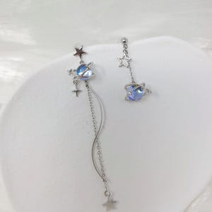 Puede incluir: Pendientes asim&eacute;tricos plateados con dise&ntilde;o celestial. Cada pendiente tiene un orbe azul similar a un planeta, estrellas y una cadena colgante. Uno tiene una cadena m&aacute;s larga con un colgante de estrella, el otro una cadena m&aacute;s corta.