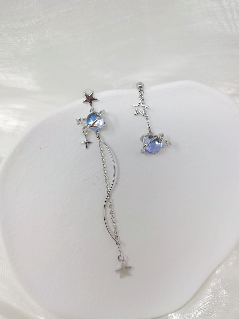 Puede incluir: Pendientes asim&eacute;tricos plateados con dise&ntilde;o celestial. Cada pendiente tiene un orbe azul similar a un planeta, estrellas y una cadena colgante. Uno tiene una cadena m&aacute;s larga con un colgante de estrella, el otro una cadena m&aacute;s corta.