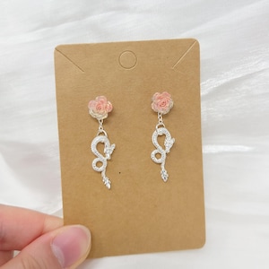 May include: A pair of dangle earrings featuring a pink rose at the top and a silver snake design. The earrings are held against a brown card.