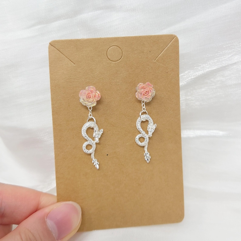 May include: A pair of dangle earrings featuring a pink rose at the top and a silver snake design. The earrings are held against a brown card.