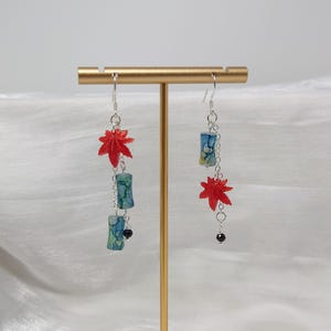 May include: Dangling earrings featuring red maple leaf charms and cylindrical beads with blue and green patterns. The earrings are suspended from silver-colored hooks and chains, with a small black bead at the bottom of one earring.