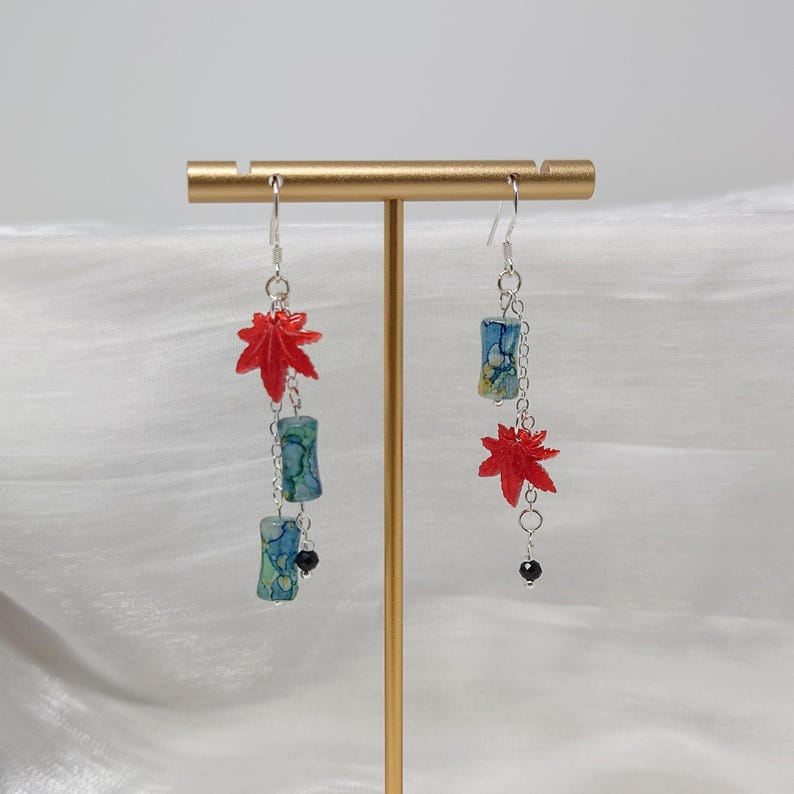 May include: Dangling earrings featuring red maple leaf charms and cylindrical beads with blue and green patterns. The earrings are suspended from silver-colored hooks and chains, with a small black bead at the bottom of one earring.