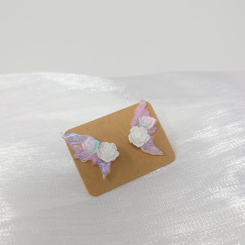 May include: A pair of handmade earrings featuring a wing design with iridescent pink and blue accents. Each wing is adorned with a white and pink rose. The earrings are displayed on a brown card.