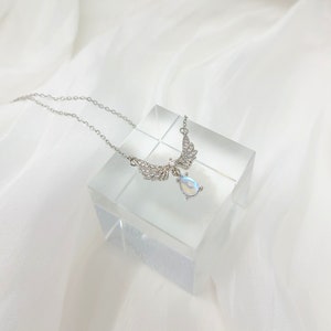 May include: A silver necklace with a teardrop-shaped opal pendant and angel wings adorned with clear crystals.