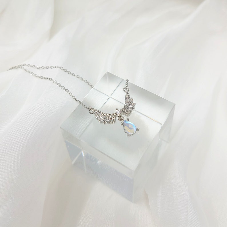 May include: A silver necklace with a teardrop-shaped opal pendant and angel wings adorned with clear crystals.