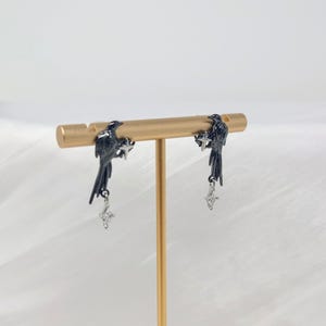 May include: A pair of black bird earrings with dangling silver-colored charms. The earrings are displayed on a gold-colored jewelry stand against a white background. The birds are perched on a bar, with the charms hanging below.