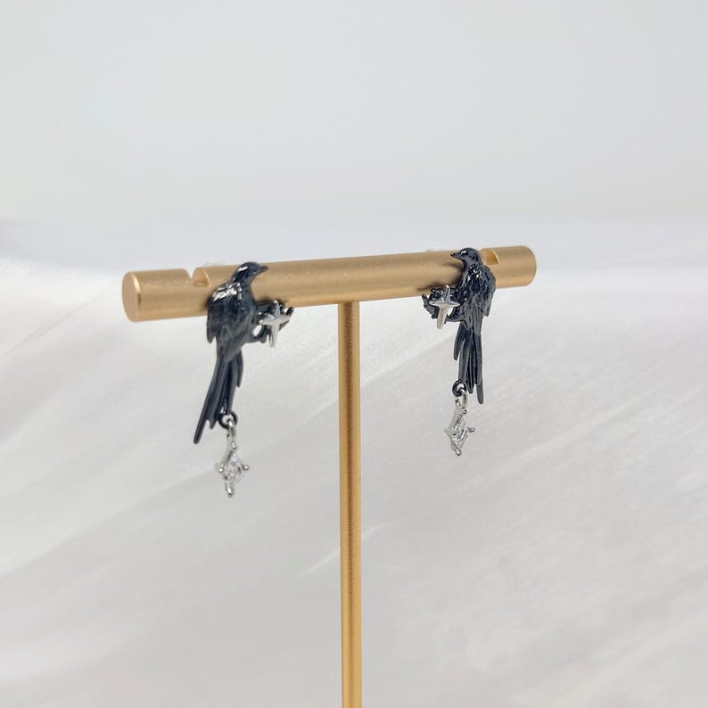 May include: A pair of black bird earrings with dangling silver-colored charms. The earrings are displayed on a gold-colored jewelry stand against a white background. The birds are perched on a bar, with the charms hanging below.
