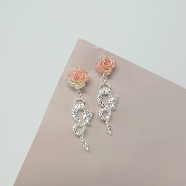 May include: A pair of dangle earrings featuring a rose stud and a silver dragon charm. The rose studs have a gradient of pink and yellow. The dragon charms are detailed with a textured design.