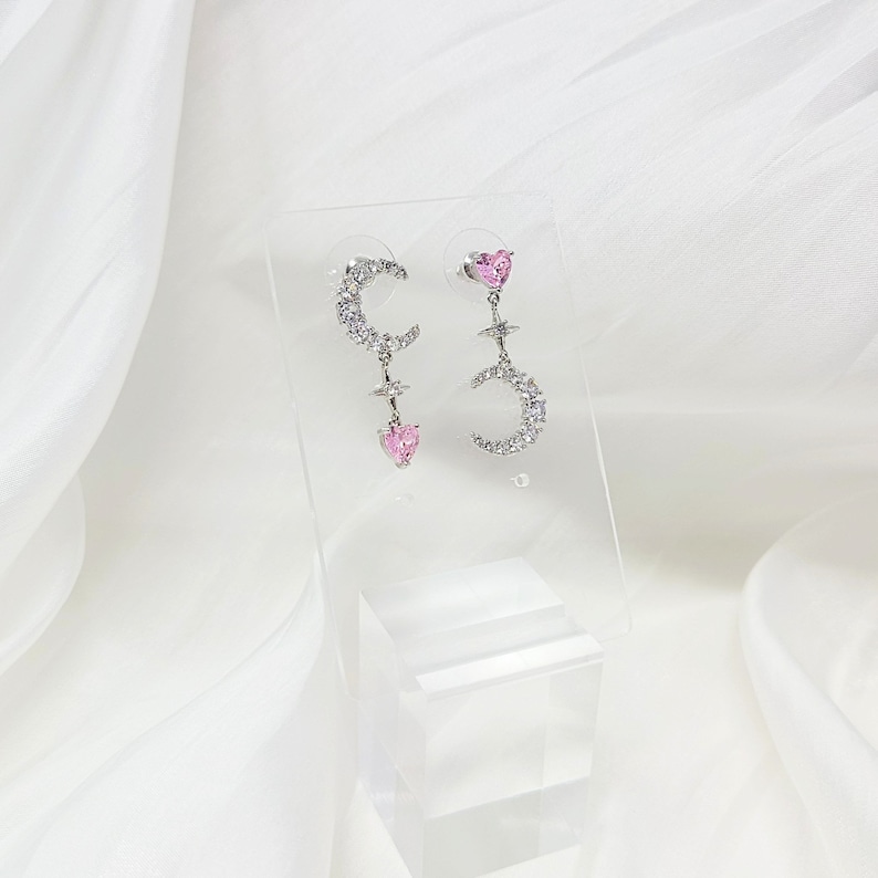May include: A pair of silver earrings with a crescent moon and star design. Each earring features a pink heart charm.