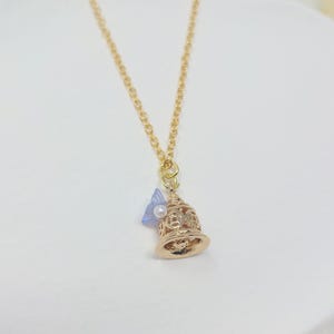 May include: A gold-colored necklace featuring a bell-shaped pendant. The pendant has intricate cut-out designs and is adorned with a small blue bow and a white pearl. The necklace chain is delicate and gold-toned.