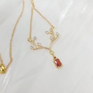 May include: A gold-colored necklace featuring a tree branch design with clear crystal accents. The pendant includes a rectangular, red gemstone. The necklace is displayed on a white, textured surface.