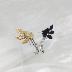 May include: A pair of decorative ear cuffs. One cuff features gold-colored wings, cream-colored roses, and silver accents. The other cuff has black wings and roses with silver accents. Both have dangling charms.