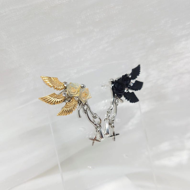 May include: A pair of decorative ear cuffs. One cuff features gold-colored wings, cream-colored roses, and silver accents. The other cuff has black wings and roses with silver accents. Both have dangling charms.
