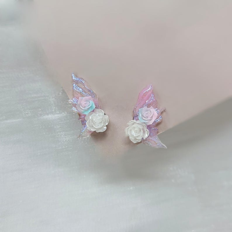 May include: A pair of delicate, fairy-inspired earrings. Each earring features a translucent, pink wing shape with iridescent glitter accents. White, light blue, and pink rose embellishments add a floral touch to the design.
