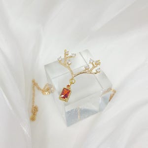 May include: A gold-colored necklace with a branch-like pendant. The pendant features small clear crystals and a rectangular, red gemstone. The necklace is displayed on a clear, square block against a white background.