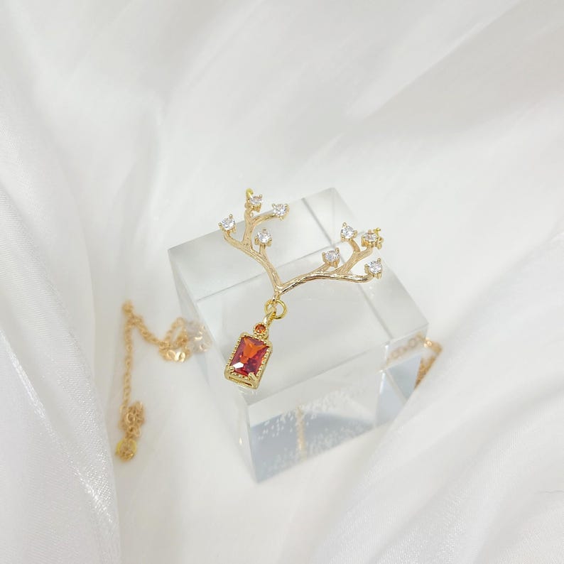 May include: A gold-colored necklace with a branch-like pendant. The pendant features small clear crystals and a rectangular, red gemstone. The necklace is displayed on a clear, square block against a white background.
