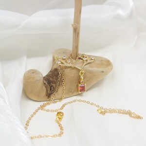 May include: A gold-colored necklace with a tree branch design. The necklace features a rectangular, red gemstone pendant and small, clear gemstones. The necklace is displayed on a piece of wood against a white background.