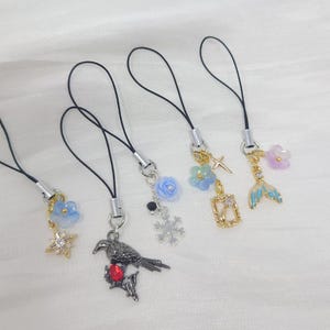 May include: Four phone charms with black cords. The first has a blue flower and gold star. The second features a dark bird with a red gem. The third has a blue rose, black bead, and snowflake. The fourth has a blue flower, cross, and frame. The fifth has a mermaid tail and purple flower.