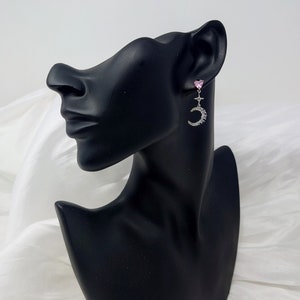 May include: A pair of silver dangle earrings with a pink heart charm and a crescent moon design. The moon is adorned with small, clear rhinestones.