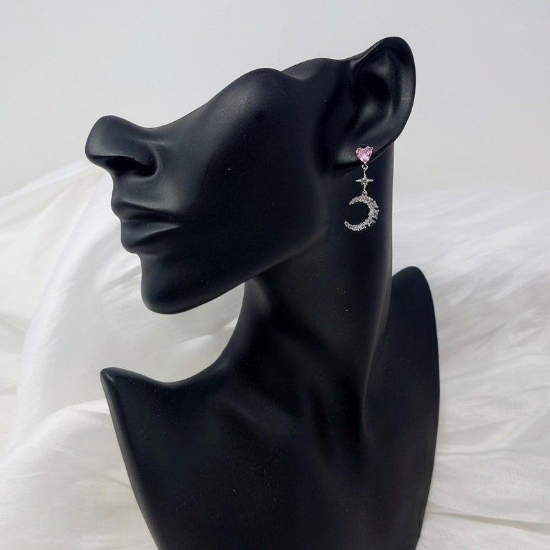 May include: A pair of silver dangle earrings with a pink heart charm and a crescent moon design. The moon is adorned with small, clear rhinestones.