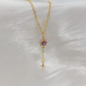 May include: A delicate gold-colored necklace featuring a pendant with a purple gemstone center surrounded by small clear stones. A small gold cross hangs below the pendant. The necklace is displayed on a white, textured surface.