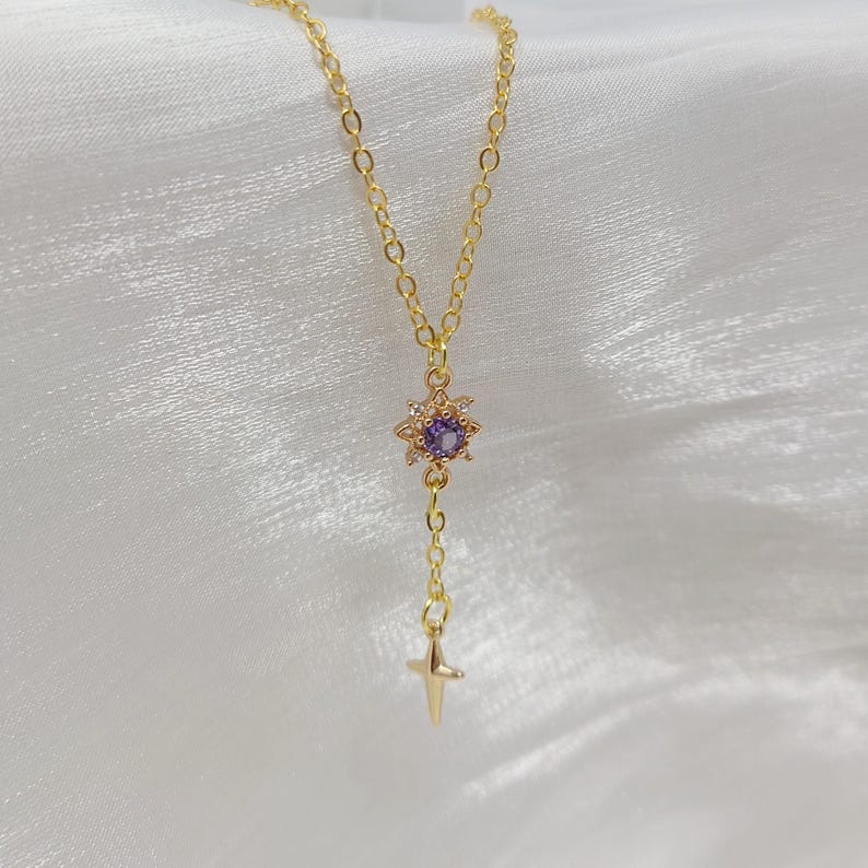 May include: A delicate gold-colored necklace featuring a pendant with a purple gemstone center surrounded by small clear stones. A small gold cross hangs below the pendant. The necklace is displayed on a white, textured surface.