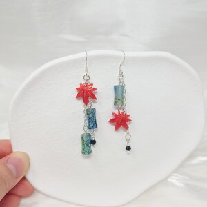 May include: Dangling earrings featuring red maple leaf charms and blue and green cylindrical beads. The earrings are connected by silver chains and have a small black bead at the bottom. The earrings are displayed on a white surface.