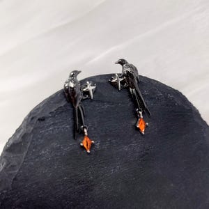 May include: A pair of black bird earrings with orange gemstone accents. The earrings feature a detailed bird design with a dangling orange gem. The earrings are set against a dark background.