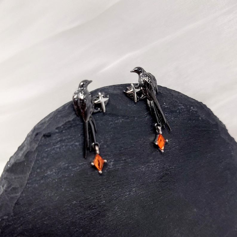 May include: A pair of black bird earrings with orange gemstone accents. The earrings feature a detailed bird design with a dangling orange gem. The earrings are set against a dark background.